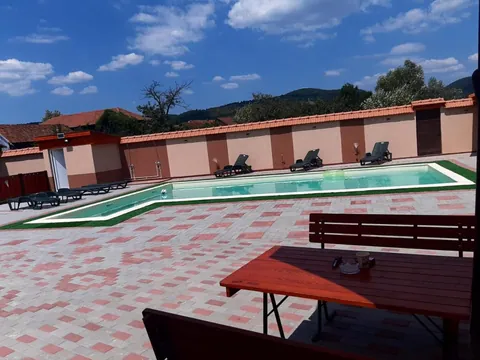 Piscina în formă de L cu marginea de gazon, mese de lemn și dealurile împădurite în fundal